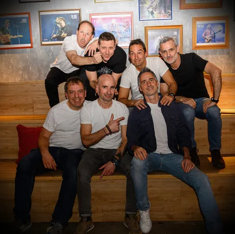 Los siete músicos posan juntos en un banco, con cuadros de música detrás.