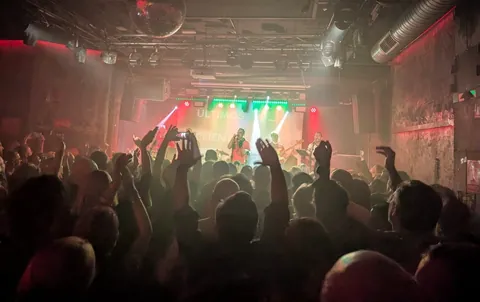 La sala llena vista desde el fondo, brazos en alto entre luces rojas y verdes.