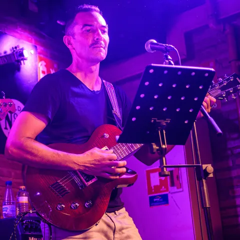 Sam toca la guitarra eléctrica junto a su micrófono, con luz azul.