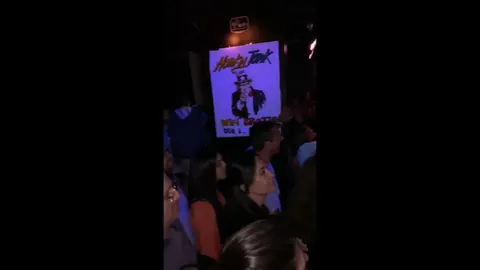El público de espaldas frente al cartel del concierto, en un bar.